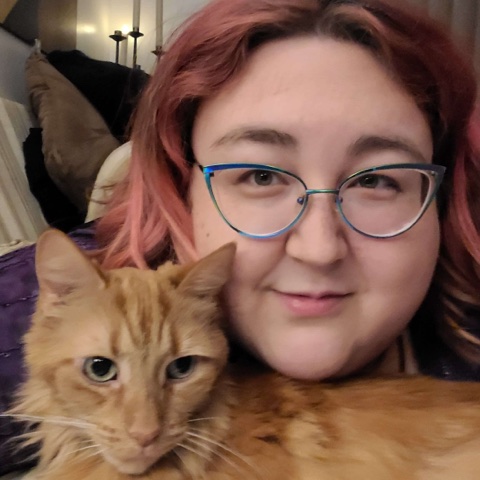 A pink haired mixed race person wearing turquoise cats eye glasses is staring at the camera with an orange cat under their chin. They are laying on their stomach on a couch and a purple shirt can be seen.