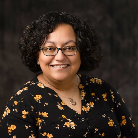 A tan Latina with short, curly, dark brown hair and glasses smiling and wearing a black dress shirt with yellow flowers.